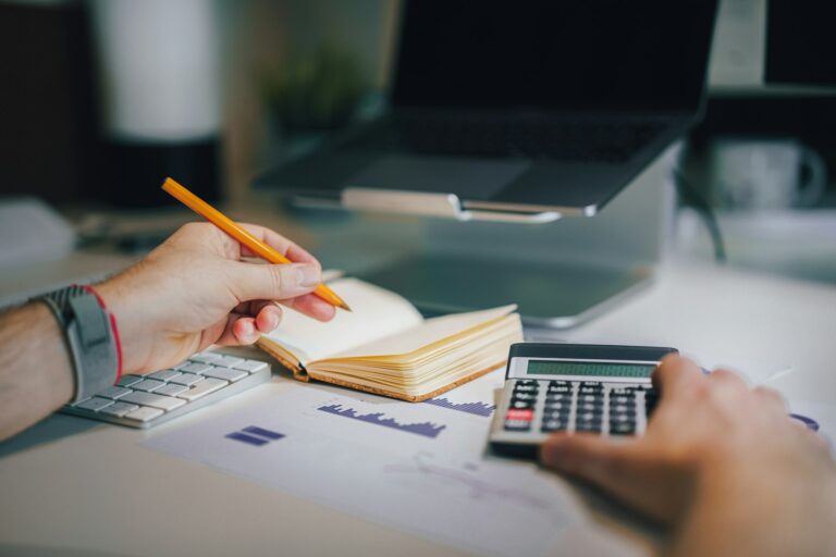 A person tracking expenses with a notebook, pencil, and calculator at a desk